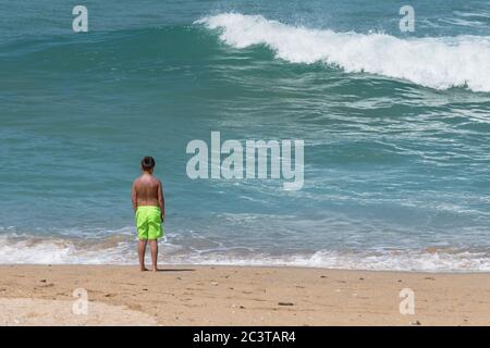 Un ragazzo giovane che indossa vibranti pantaloncini verdi calce che si levano in piedi da solo sulla costa guardando le onde a Fistral a Newquay in Cornovaglia. Foto Stock