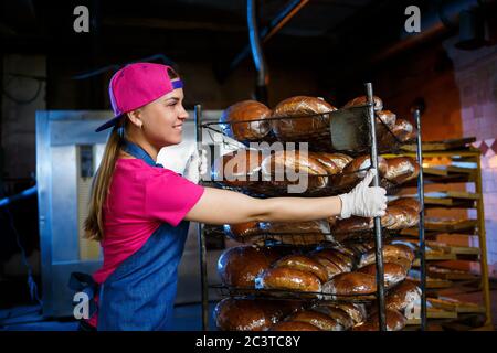 Una ragazza panettiera prende il pane caldo in una panetteria sullo sfondo di scaffalatura con il pane. Produzione industriale di pane. La fase di cottura in un panettiere Foto Stock