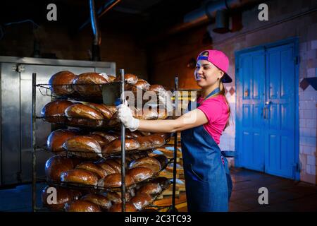 Una ragazza panettiera prende il pane caldo in una panetteria sullo sfondo di scaffalatura con il pane. Produzione industriale di pane. La fase di cottura in un panettiere Foto Stock