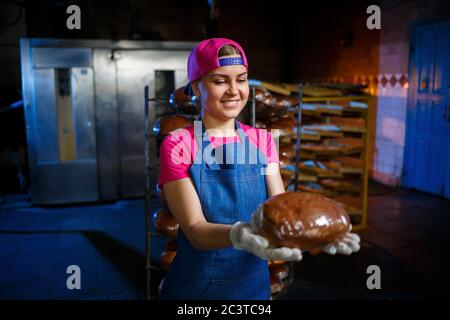 Una ragazza panettiera prende il pane caldo in una panetteria sullo sfondo di scaffalatura con il pane. Produzione industriale di pane. La fase di cottura in un panettiere Foto Stock