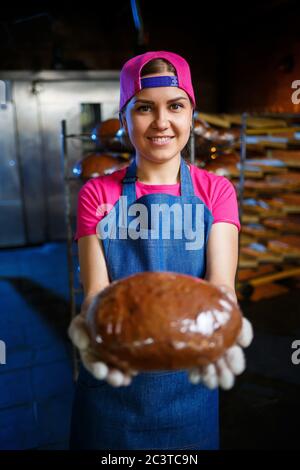 Una ragazza panettiera prende il pane caldo in una panetteria sullo sfondo di scaffalatura con il pane. Produzione industriale di pane. La fase di cottura in un panettiere Foto Stock