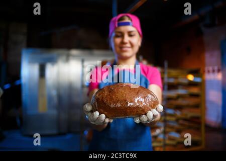 Una ragazza panettiera prende il pane caldo in una panetteria sullo sfondo di scaffalatura con il pane. Produzione industriale di pane. La fase di cottura in un panettiere Foto Stock