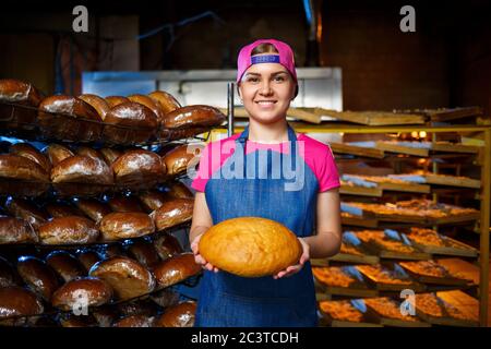 Una ragazza panettiera prende il pane caldo in una panetteria sullo sfondo di scaffalatura con il pane. Produzione industriale di pane. La fase di cottura in un panettiere Foto Stock