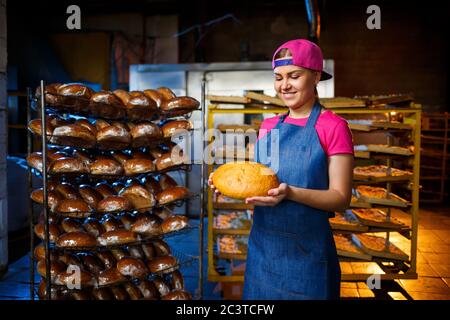 Una ragazza panettiera prende il pane caldo in una panetteria sullo sfondo di scaffalatura con il pane. Produzione industriale di pane. La fase di cottura in un panettiere Foto Stock