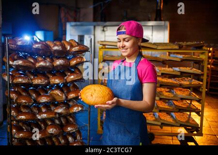 Una ragazza panettiera prende il pane caldo in una panetteria sullo sfondo di scaffalatura con il pane. Produzione industriale di pane. La fase di cottura in un panettiere Foto Stock