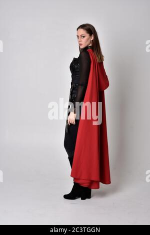 Ritratto di bruna ragazza che indossa tuta in pelle nera e mantello rosso. Posa in piedi a lunghezza intera, isolata contro uno sfondo di studio. Foto Stock