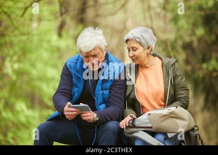 Ritratto di coppia senior attiva utilizzando smartphone mentre si riposa durante l'escursione nella foresta autunnale, copia spazio Foto Stock