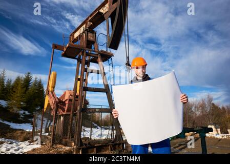 Ritratto di un operaio in uniforme blu e casco arancione in piedi in un campo petrolifero con un disegno stampato in una bella giornata di sole, olio pompa jack e cielo blu su sfondo Foto Stock