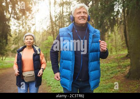 Ritratto della coppia senior attiva che corre nella foresta, concentrarsi su un uomo sorridente indossando le cuffie e ascoltando musica Foto Stock