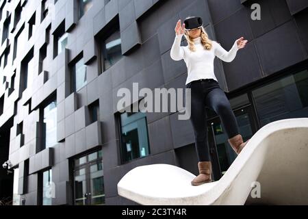Donna bionda che indossa occhiali di realtà virtuale contro l'edificio futuristico Foto Stock