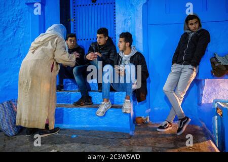 Marocco: Chefchaouen, la città blu. Atmosfera nella Città Vecchia: Vecchia donna in abiti tradizionali che parla con giovani uomini seduti su una scala in un l Foto Stock