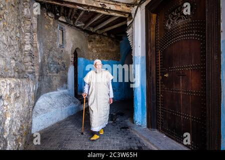 Marocco, Tangeri: Atmosfera in una strada della Città Vecchia. Uomo anziano che indossa abiti tradizionali camminando con un bastone, passando da una porta finemente lavorata Foto Stock