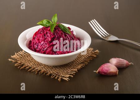 Insalata di barbabietola bollita grattugiata con aglio e maionese in un recipiente bianco su un tavolo marrone. Contorno e contorno vegetariano. Cucina russa. Vista frontale. Foto Stock