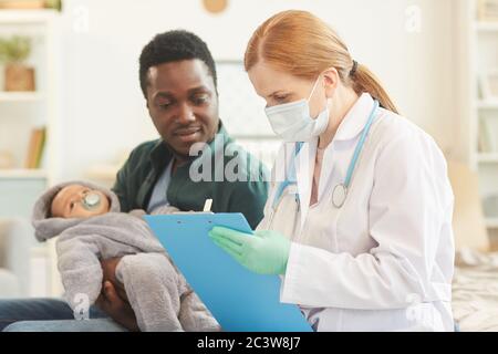 Vista laterale ritratto di donna medico scrittura su appunti durante la visita a casa per il controllo del bambino, spazio di copia Foto Stock