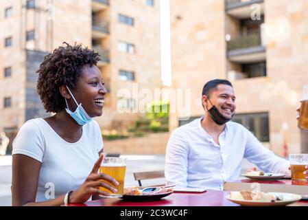 un gruppo di persone multietniche che bevono una birra, parlano e ridono in modo protettivo, socialmente distanziando e bevono in modo sicuro, indossando una maschera sotto il mento Foto Stock