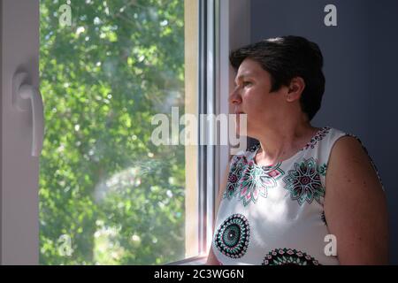 Donna matura triste, preoccupata e premurosa che guarda attraverso la finestra. Tristezza e concetto di quarantena. Foto Stock