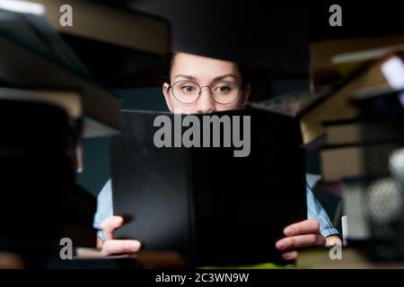 La ragazza in occhiali sta leggendo un libro. Lo studente è disseminato di libri Foto Stock