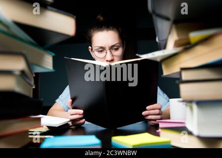 Una giovane donna con occhiali legge attentamente un libro in ritardo di notte al tavolo Foto Stock