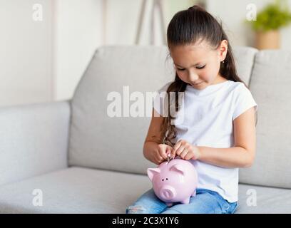 Ragazza che mette il moneta in Piggybank seduta sul divano a casa Foto Stock