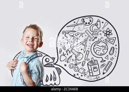 Un bambino felice in una maglietta di luce va anche a scuola per la prima volta. Dietro di lui è uno zaino in cui una varietà di icone si torna a scuola Foto Stock