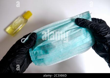 Due mani in guanti neri che tengono una maschera facciale su uno sfondo bianco. Igienizzatore per le mani giallo. Diffusione di virus e prevenzione dei batteri. Misure di sicurezza Foto Stock