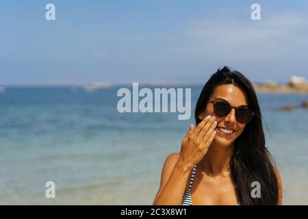 Donna sorridente che applica crema solare sul viso in spiaggia Foto Stock