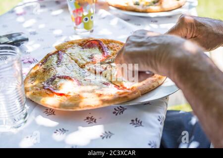 Un uomo anziano taglia una fetta di pizza salame con pepperoni su un piatto, Germania Foto Stock