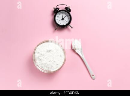 Polvere di collagene in una ciotola con un cucchiaino da tè e un orologio su sfondo rosa. Vista dall'alto. Foto Stock