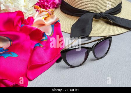 Occhiali da sole, cappello di paglia, sciarpa di seta fucsia, peonie bianche e rosate su sfondo lilla. Primo piano, piatto Foto Stock