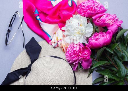 Mescolare bouquet di peony, cappello di paglia femminile, occhiali da sole e zattera di seta su sfondo lilla. Vista dall'alto. Idea per poster, sfondo Foto Stock