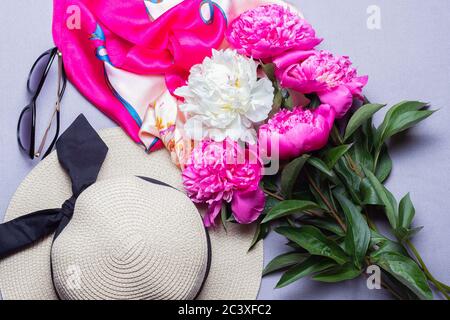 Vista dall'alto del cappello di paglia con nastro nero, bouquet di pony bianco e fucsia, sciarpa di seta e occhiali da sole isolati su sfondo lilla. Disposizione piatta, primo piano. Foto Stock