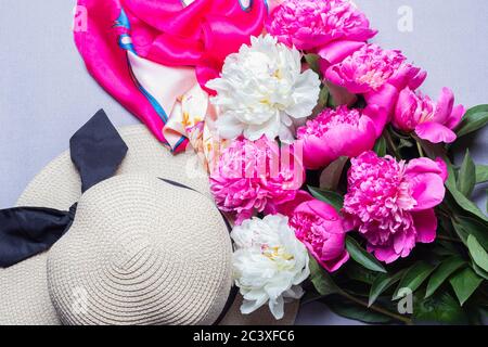 Bouquet di fucsia e peonie bianche, sciarpa di seta e cappello di paglia femminile su sfondo lilla. Vista dall'alto. Idea per poster, sfondo Foto Stock