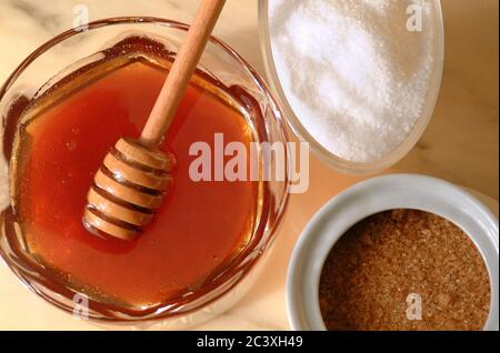 Primo piano in un angolo di posa piatto di un vaso di miele accanto a zucchero bianco e scodelle di zucchero di canna. Foto Stock