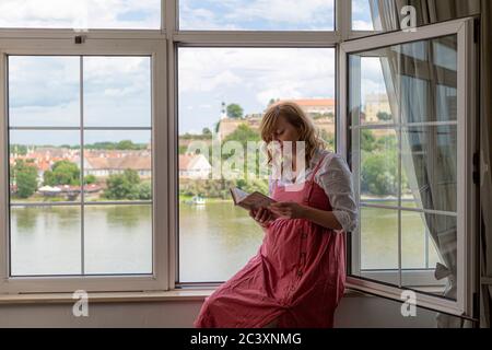 Una donna caucasica sorridente incinta seduta accanto alla finestra mentre legge un libro e la parte della città vecchia e del fiume sono sullo sfondo. Foto Stock