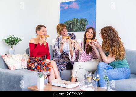 Quattro amici sorridenti che chiacchierano e si siedono sul divano nel soggiorno mentre mangiano la pizza, ci sono anche bicchieri con vino sul tavolo. Foto Stock