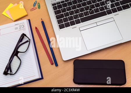 Vista dall'alto della scrivania dell'ufficio domestico: Computer portatile, smartphone, note adesive, calendario, occhiali, clip, matite colorate Foto Stock