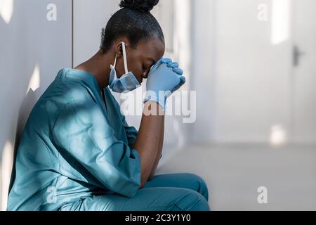 Stanco depresso femmina africana infermiera indossando maschera si siede sul pavimento dell'ospedale. Foto Stock