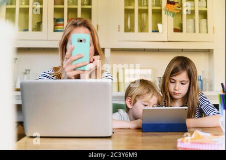 Donna che tiene lo smartphone davanti al computer portatile mentre è ragazzo (4-5) e ragazza (6-7) che utilizza un tablet digitale Foto Stock