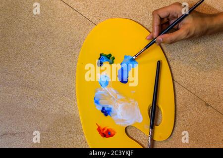 Tavolozza di pittura ad olio piena di colori e pennelli tenuti sul pavimento. La mano con un pennello mescola i colori Foto Stock