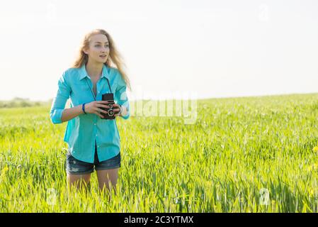 giovane donna bionda con macchina fotografica vintage su un campo di grano verde estivo che sente l'ispirazione e scatta le foto Foto Stock