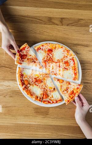 Due mani da donna rubano fette di pizza margherita da una tavola rotonda in legno, primo piano vista dall'alto foto Foto Stock