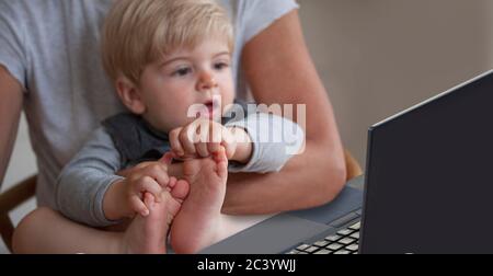 Piccolo bambino che guarda eccitato e sorpreso di qualcosa su un monitor di computer portatile in grembo di madre. Messa a fuoco selettiva sui piedi del bambino. Foto Stock