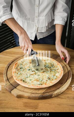 Tagliando la pizza con un taglierina, servita su tavola rotonda in legno, luce naturale Foto Stock