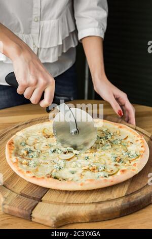 Tagliando la pizza con un taglierina, servita su tavola rotonda in legno, luce naturale Foto Stock