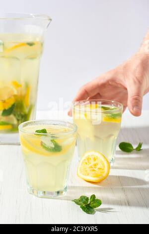 Due bicchieri e una caraffa di limonata sul tavolo bianco. Una mano dell'uomo che raggiunge per un bicchiere. Foto Stock