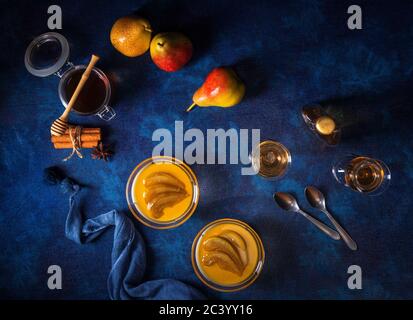 Panna cotta dessert tradizionale italiano con pere in camicia al rum e sciroppo di miele. Tavola di sfondo blu scuro, bicchieri di rum, vasetto di miele, pere e. Foto Stock