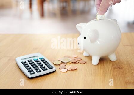 Mano sta mettendo un euro in una banca bianca del piggy su un tavolo con più monete e un calcolatore, concetto di economia per calcolare le finanze e salvare househol Foto Stock