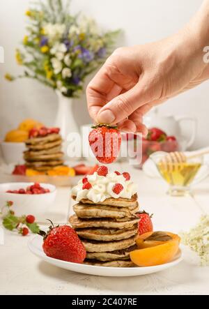 Pancake con fragole, deliziosa colazione. Una donna decora le fragole con una pila di frittelle con panna montata. Instagram verticale cibo p Foto Stock