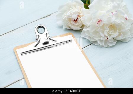 Clipboard foglio bianco di carta e fiori di pony su tavolo di legno blu. Disposizione piatta, vista dall'alto. Wedding planner, checklist, per fare la lista concetto. Foto Stock