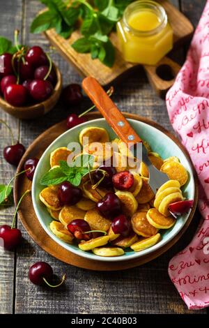 Colazione con minuscoli pancake. Mini frittelle con ciliegie e miele su rustico tavolo di legno. Foto Stock